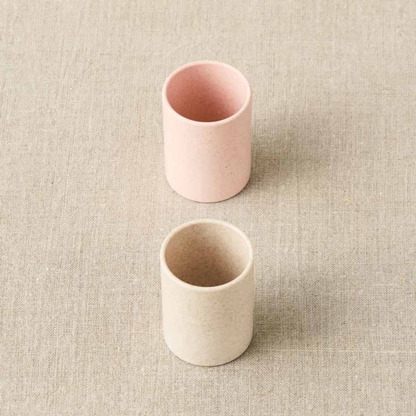 Two ceramic cups, one pink and one beige, on a textured beige surface.