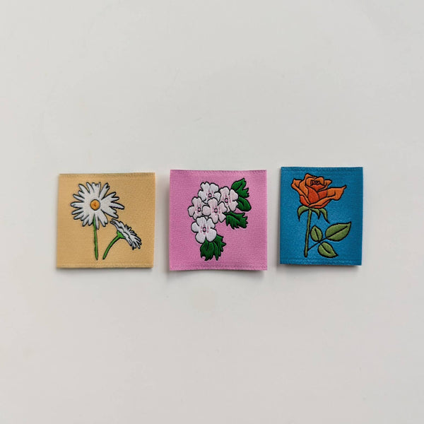 Three small square embroidered patches with floral designs, arranged horizontally on a light gray background. The left patch is beige with a white daisy, the middle is pink with white flowers, and the right is blue with an orange rose.