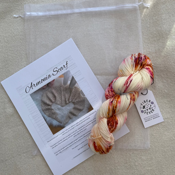 A skein of variegated yarn with a white tag reading 'Armenia Scarf' is arranged on a beige fabric background alongside a clear zip pouch, a printed card titled 'Armenia Scarf,' and a small circular logo tag. The yarn has cream, peach, orange, and red variegated tones.