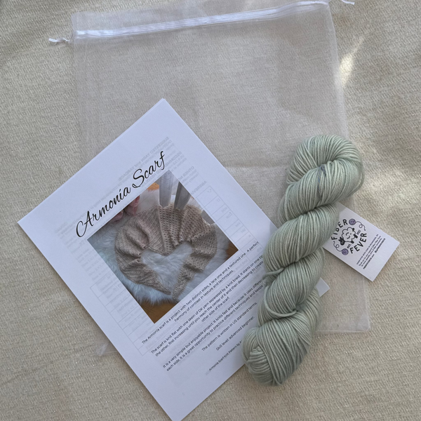 A skein of pale blue/green yarn next to a clear drawstring pouch containing a printed card labeled 'Ammonia Scarf' and a small product tag, arranged on a beige fabric background.