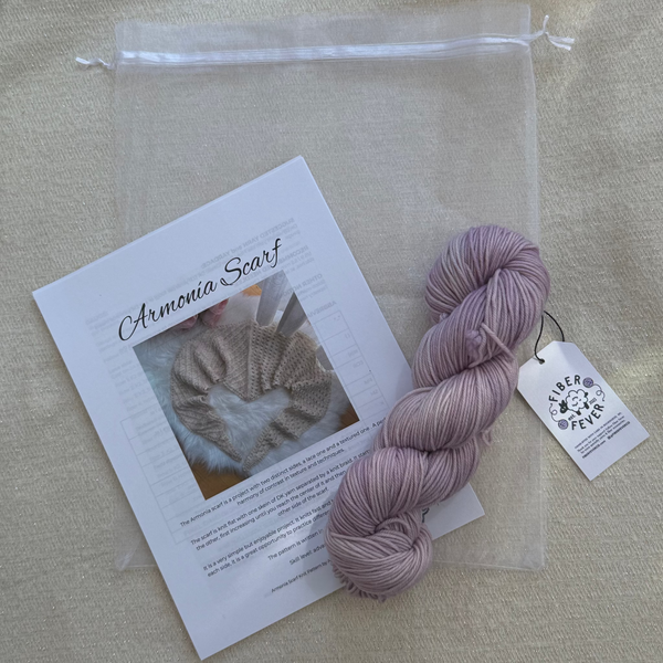 A skein of dusty lavender yarn next to a clear drawstring bag and an informational card titled 'Armonia Scarf' featuring a cat photo, all arranged on a beige fabric background.