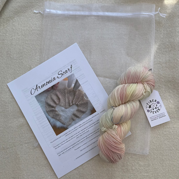 A skein of variegated yarn in soft pastel shades of pink, cream, and pale yellow, displayed next to a clear drawstring bag and a printed card labeled 'Artemia Scarf' on a beige fabric background.
