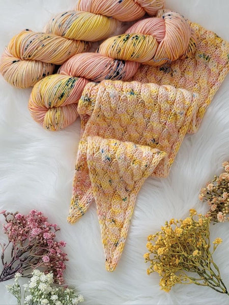 A scarf kit displayed with a knitted scarf in pink and yellow hues, alongside skeins of yarn in a matching color palette, and dried flowers around it.