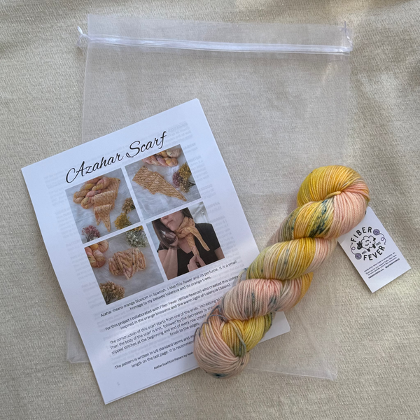 A skein of variegated yarn in soft pastel shades of yellow, peach, pink, and green, displayed next to a printed instruction card labeled 'Cznhar Scarf' and a clear zip bag on a beige fabric background.