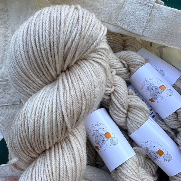 Cream-colored yarn skeins in a natural canvas bag, with white paper labels featuring a cartoon bear and the brand name 'The Knit Life'.
