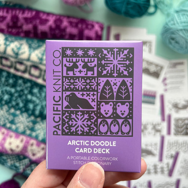 Hand holding a purple card deck with yarn and knitting patterns in the background