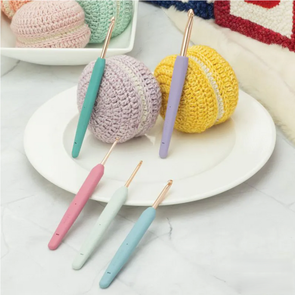 Four crochet hooks with pastel-colored handles in pink, mint, light blue, and lavender are arranged on a white plate with matching crocheted ball toppers in pastel tones.