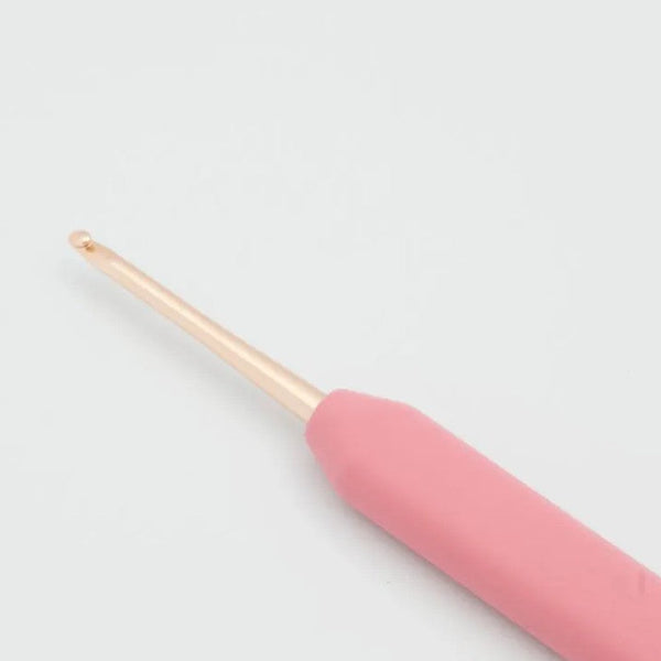 A close-up of a crochet hook with a pink ergonomic handle and a metallic rose gold tip, set against a plain light gray background.